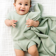 Baby in a green outfit lying on a white textured surface with a green blanket.