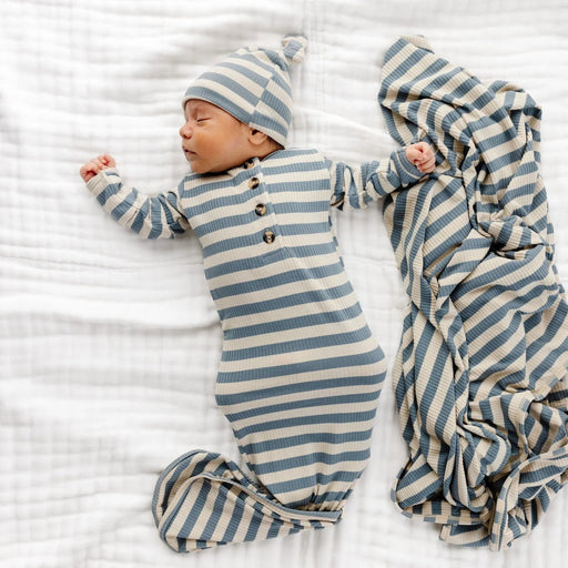 A newborn baby lying on a white muslin blanket, wearing a matching blue and cream striped footed sleeper and hat, with a folded striped swaddle blanket beside them.