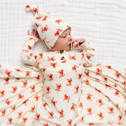 Baby swaddled in a white blanket with red patterns, wearing a matching hat on a white background