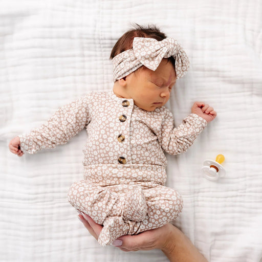 Newborn baby in a floral outfit with a headband, lying on a white textured blanket.