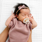 Newborn baby in a pink knitted outfit with a pacifier, lying on a white textured surface.