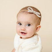 Baby wearing a white outfit and headband with a bow against a beige background