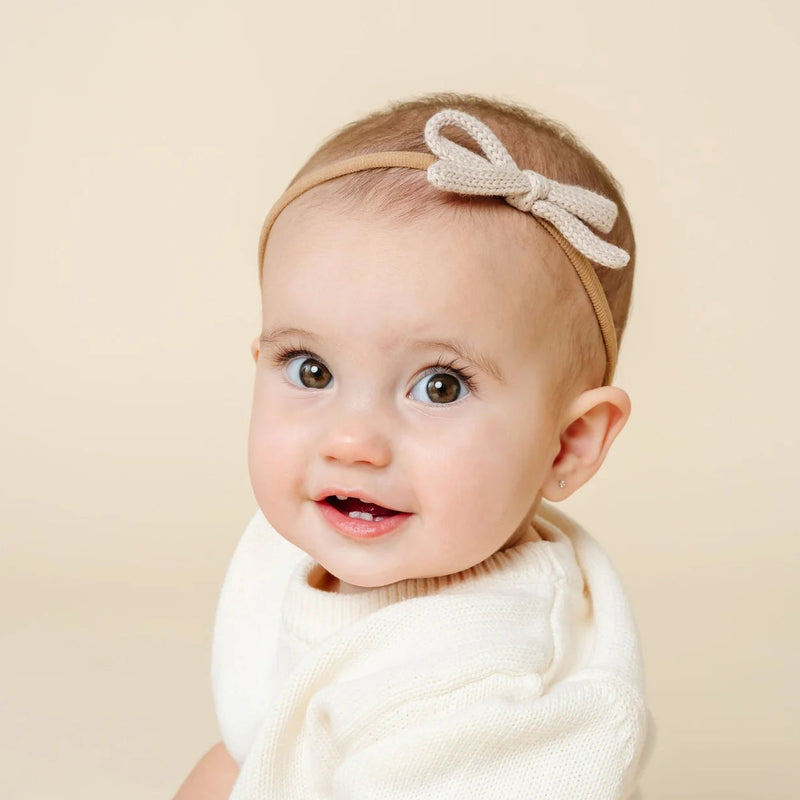 Baby wearing a white outfit and headband with a bow against a beige background