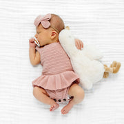 A newborn baby lying on a white textured blanket, wearing a dusty pink ribbed romper with a ruffled hem, a matching pink bow headband, and holding a white plush toy.