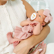 A baby wearing a pink knit romper and matching pink bow headband rests on a woman's lap, with a pacifier in their mouth. The woman wears a white knit cardigan.
