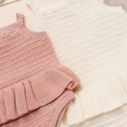 A styled flat-lay of baby knit romper sets on a warm beige background. The set in the foreground is dusty pink with a ruffled hem and three decorative buttons.