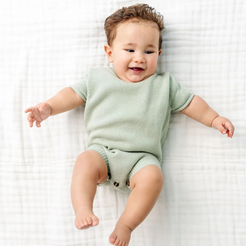 Baby wearing a green knit sweater outfit lying on a white textured surface