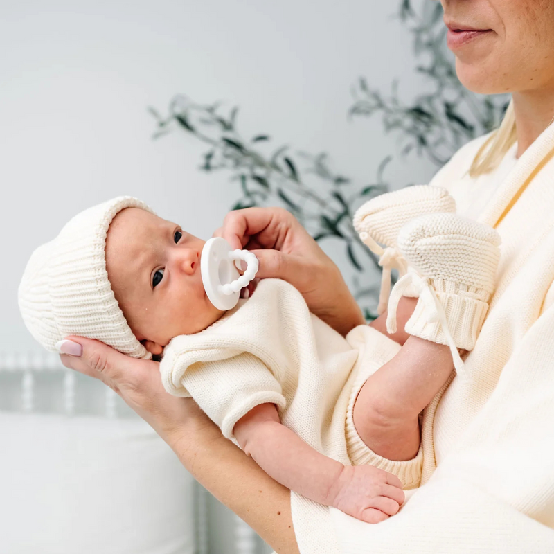Baby in a white outfit and hat held by an adult, with a neutral background