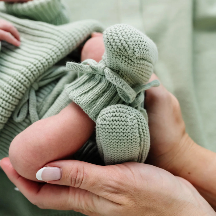Green knitted baby booties held in a hand against a neutral background
