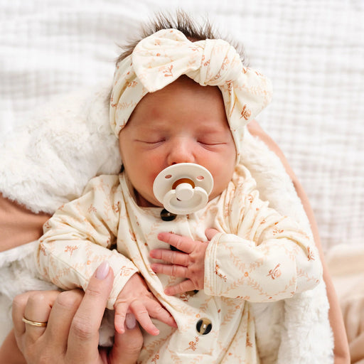 Newborn baby in Lou Lou & Co Annie floral gown and matching headband sleeping in parent’s arms with neutral blanket