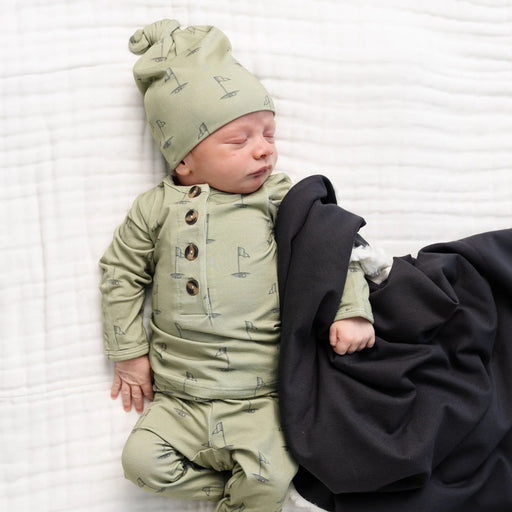 Newborn baby wearing Lou Lou & Co Cal print top and bottom set with matching hat in light green golf flag pattern, lying on a white blanket