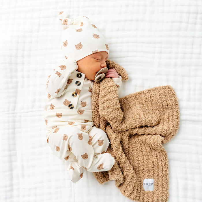 Newborn baby wearing Lou Lou & Co Teddy bear print top and bottom set with matching hat and pacifier lying on white blanket with brown knit blanket