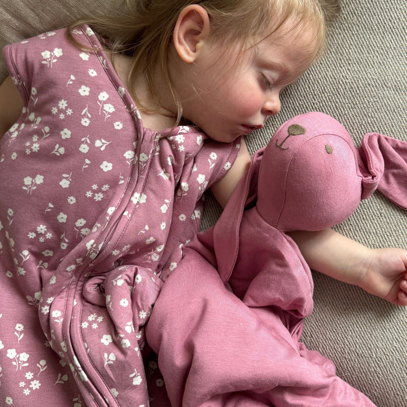 A toddler girl sleeps peacefully on a beige couch while hugging a pink Bunny Lovey and wearing a pink mini floral günamüna sleep sack. The lovey has long floppy ears and a soft blanket-style body.