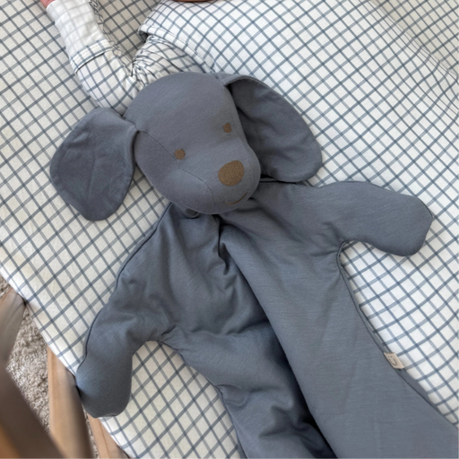 A baby is sleeping peacefully in a crib on a checkered sheet, holding a soft gray dog-shaped lovey with floppy ears and an elongated blanket body.