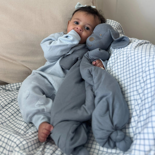 A baby sits on a bed leaning against pillows while holding the gray Pup Lovey upright, looking at it with curiosity.