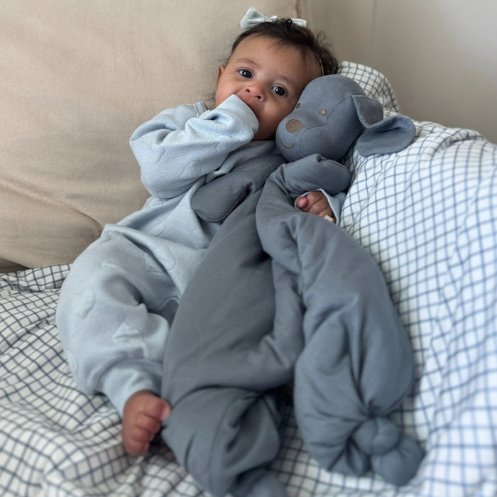 A baby sits on a bed leaning against pillows while holding the gray Pup Lovey upright, looking at it with curiosity.