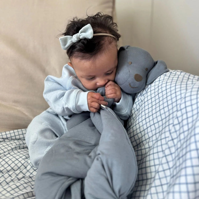 A baby sits on a bed hugging the gray Pup Lovey close to their face while smiling, with neutral pillows behind them.