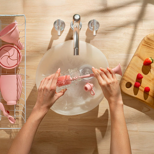 Person cleaning a bottle with a brush in a sink, with pink kitchen utensils and a wooden cutting board with strawberries in the background.