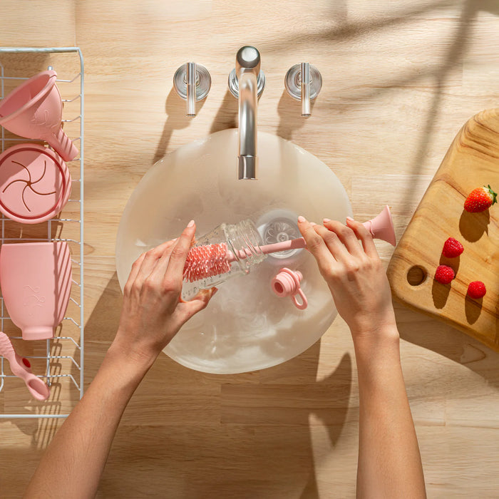 Person cleaning a bottle with a brush in a sink, with pink kitchen utensils and a wooden cutting board with strawberries in the background.