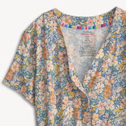 A close-up of a short-sleeve button-front top with a blue floral print, featuring white, peach, and yellow flowers with green leaves. The inner collar is decorated with a colorful striped band, and the inside label shows the brand 'You're Welcome'.