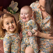 A mother and daughter wearing matching floral print baby jumpsuits, with the mother holding a smiling baby in a cozy indoor setting.
