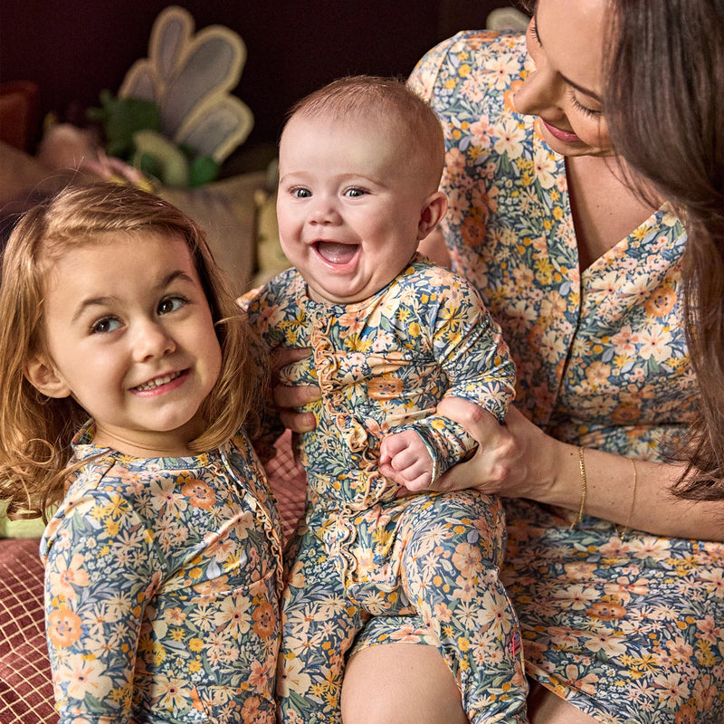 A mother and daughter wearing matching floral print baby jumpsuits, with the mother holding a smiling baby in a cozy indoor setting.