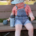 Child sitting on a bench with a water bottle and snack cup, wearing a pink shirt and denim overalls.