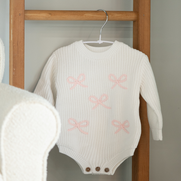 White baby romper with pink bows on a wooden hanger against a gray wall.
