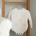 White baby romper with pink bows on a wooden hanger against a gray wall.