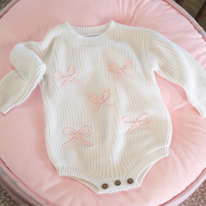 White knitted baby romper with pink bows on a pink cushion