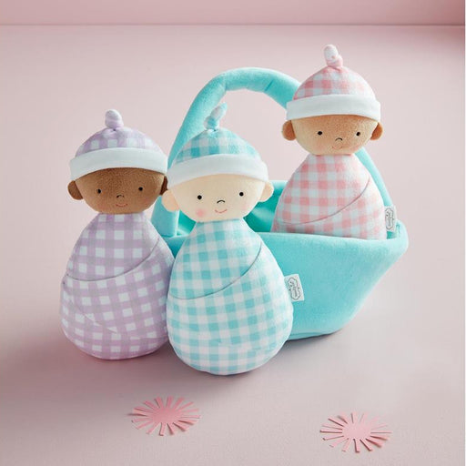Three soft plush baby dolls wrapped in pastel gingham blankets and hats, with one doll sitting in a light blue basket on a pink studio background.