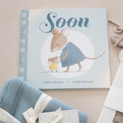 Children's book titled 'Soon' with baby booties and blankets on a beige background