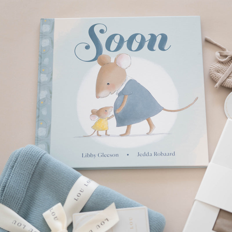 Children's book titled 'Soon' with baby booties and blankets on a beige background