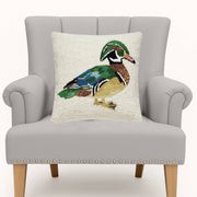A square throw pillow featuring a detailed, textured needlepoint-style wood duck design on an off-white background, displayed on a light gray upholstered armchair.