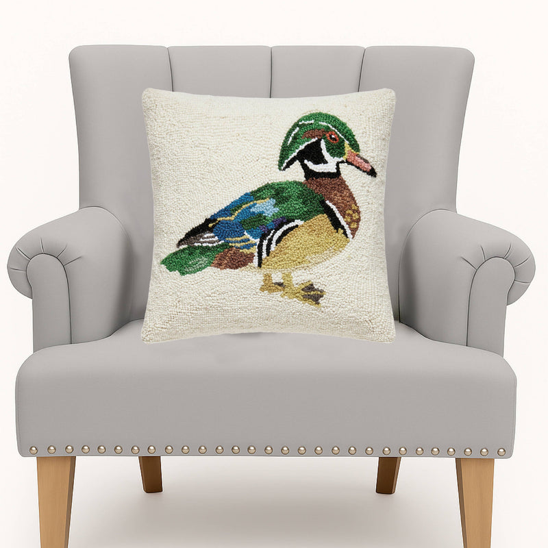 A square throw pillow featuring a detailed, textured needlepoint-style wood duck design on an off-white background, displayed on a light gray upholstered armchair.