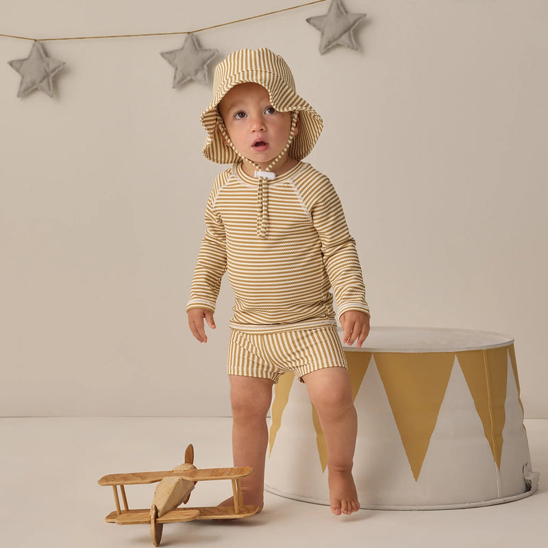 Baby in striped outfit and sun hat standing next to a wooden airplane and white drum with star decorations on the wall.