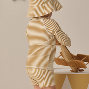 Child wearing a yellow and white striped outfit with a matching hat, standing next to a wooden toy.