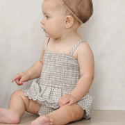 Baby wearing a checkered romper sitting on a wooden floor against a plain background