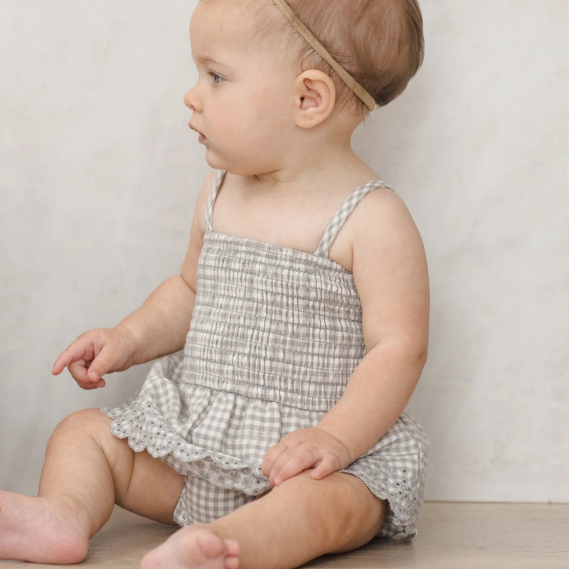 Baby wearing a checkered romper sitting on a wooden floor against a plain background