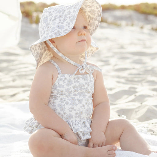Baby in a floral swimsuit and sun hat sitting on sand at the beach