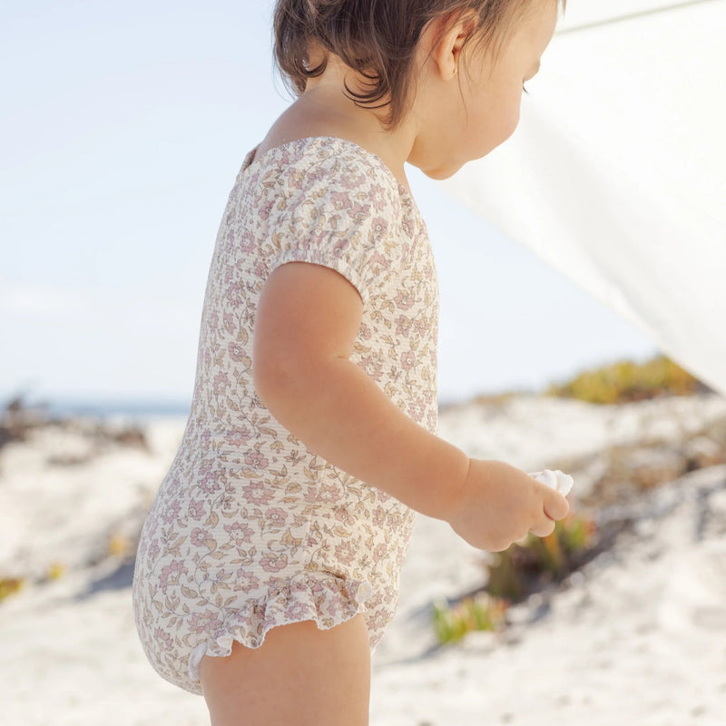 Child in a floral one piece swimsuit standing on a sandy beach with a blurred background