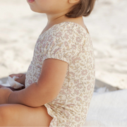 Child wearing a floral dress sitting on a sandy beach.