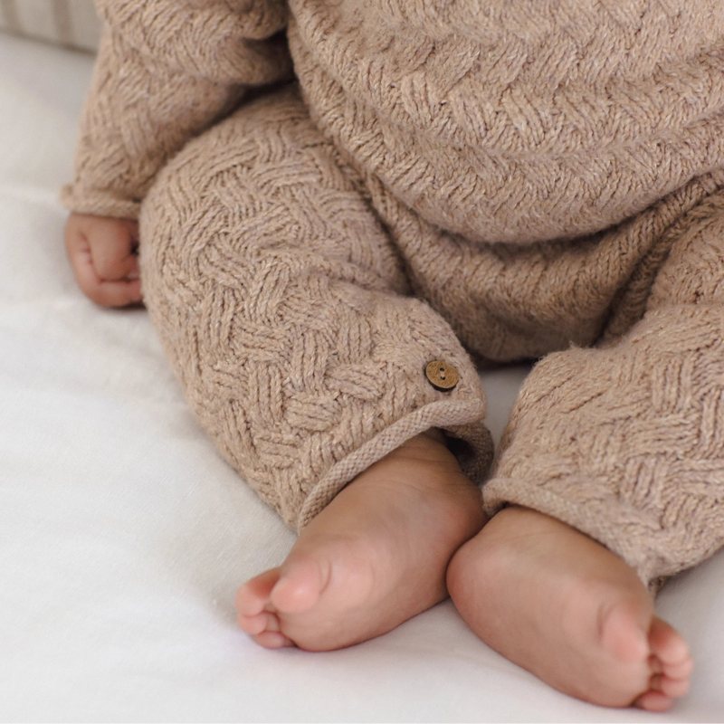 Close-up of a baby wearing a chunky knit beige jumpsuit with a textured cable pattern and a small wooden button detail on the cuff, sitting on a white blanket.