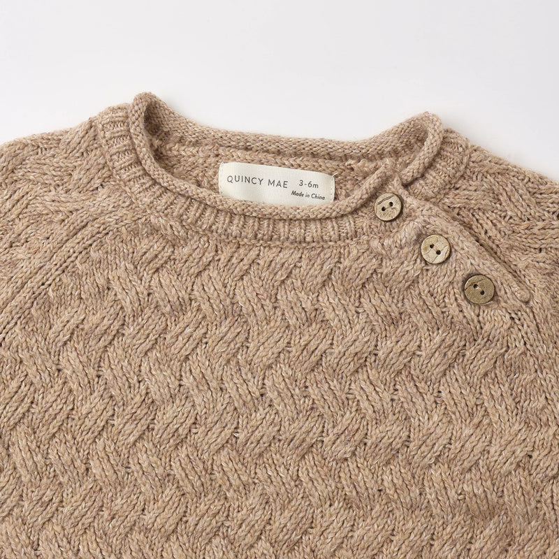Close-up of a beige cable-knit baby sweater with a mock neck, three-quarter button closure, and a visible Quincy Mae brand label.