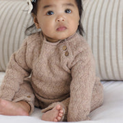 A baby sitting on a white bed wearing a beige cable-knit long-sleeve romper with brown buttons, set against a background of striped beige pillows.