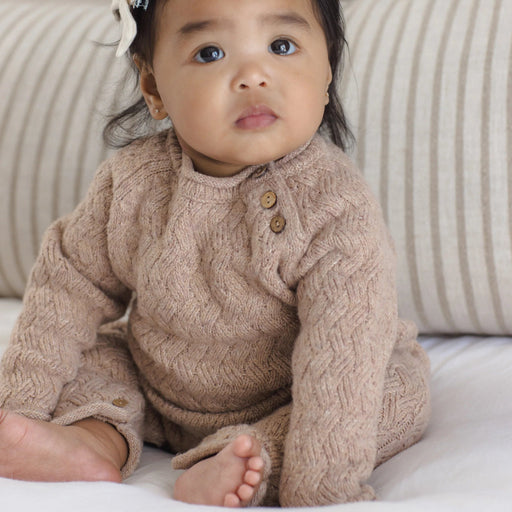 A baby sitting on a white bed wearing a beige cable-knit long-sleeve romper with brown buttons, set against a background of striped beige pillows.
