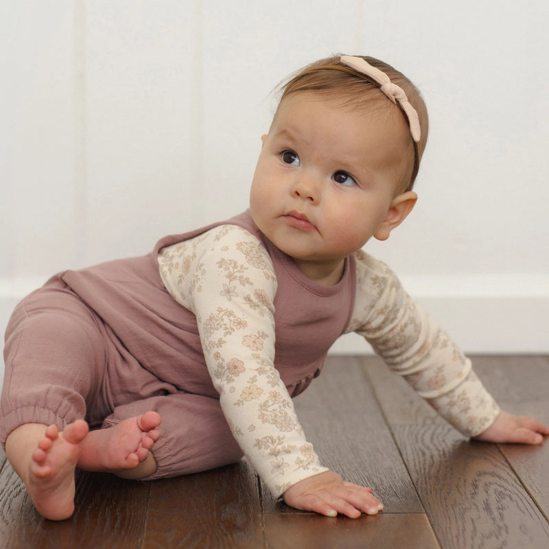 A baby crawling on a dark wood floor against a white paneled wall, wearing a mauve-brown sleeveless jumpsuit over a cream floral long-sleeve shirt and a matching pink headband.
