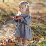 A young toddler girl stands on a leaf-covered path outdoors, wearing a dusty blue-gray long-sleeve dress with front pockets and holding red apples. A small basket with apples sits beside her.