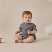 Baby wearing a gray knitted outfit sitting on a beige floor with a neutral background
