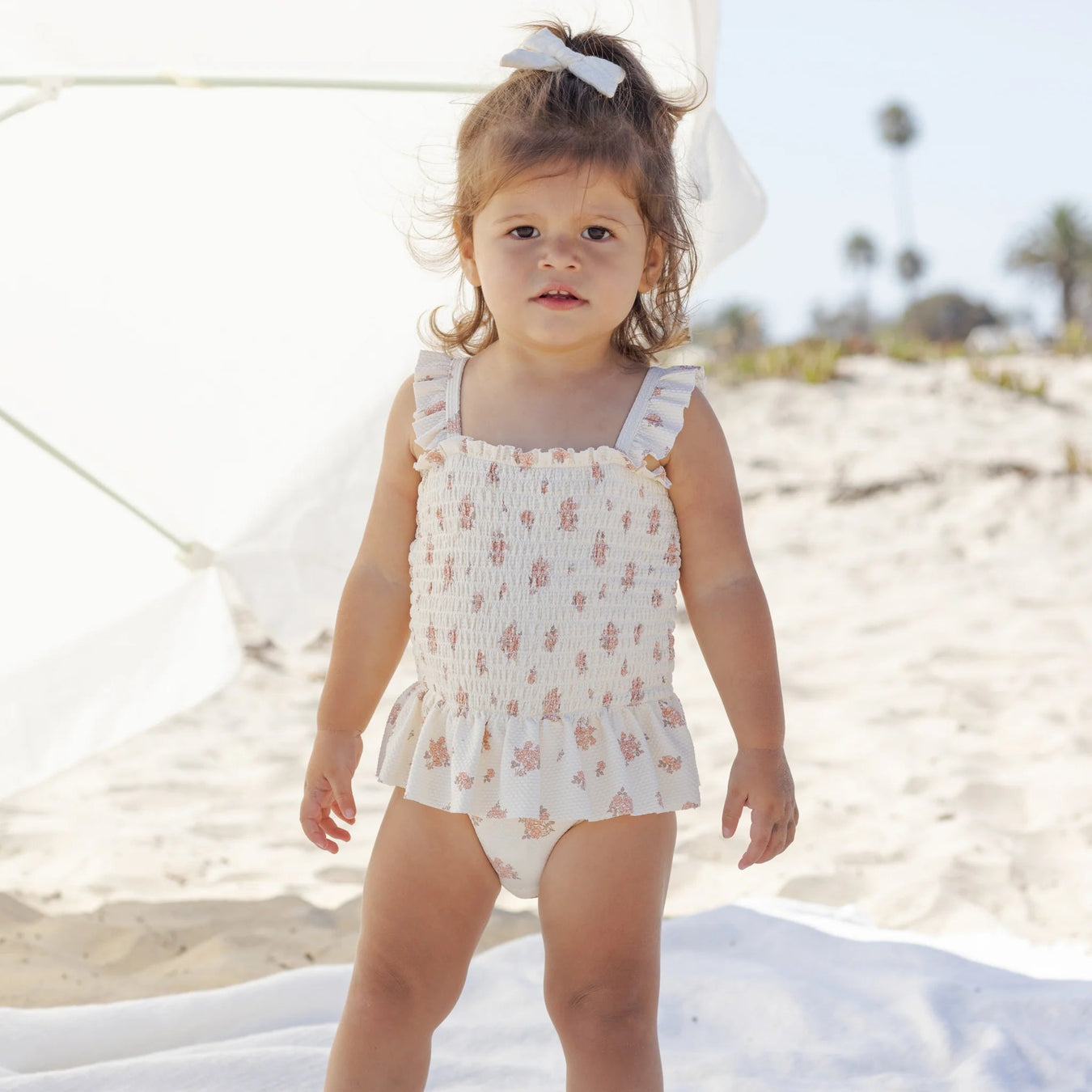 Young child in a floral swimsuit standing on a sandy beach.